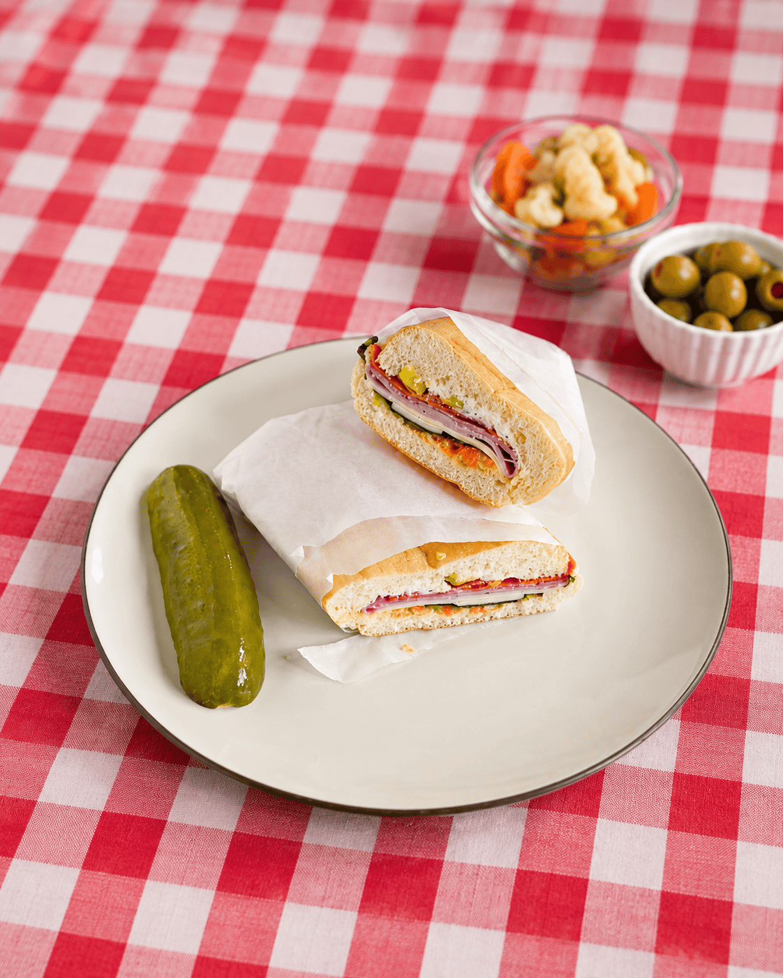 A plate with 2 sandwich halves stacked on top of each other and a pickle with a bowl of olives and a bowl of pickled veggies behind the plate.