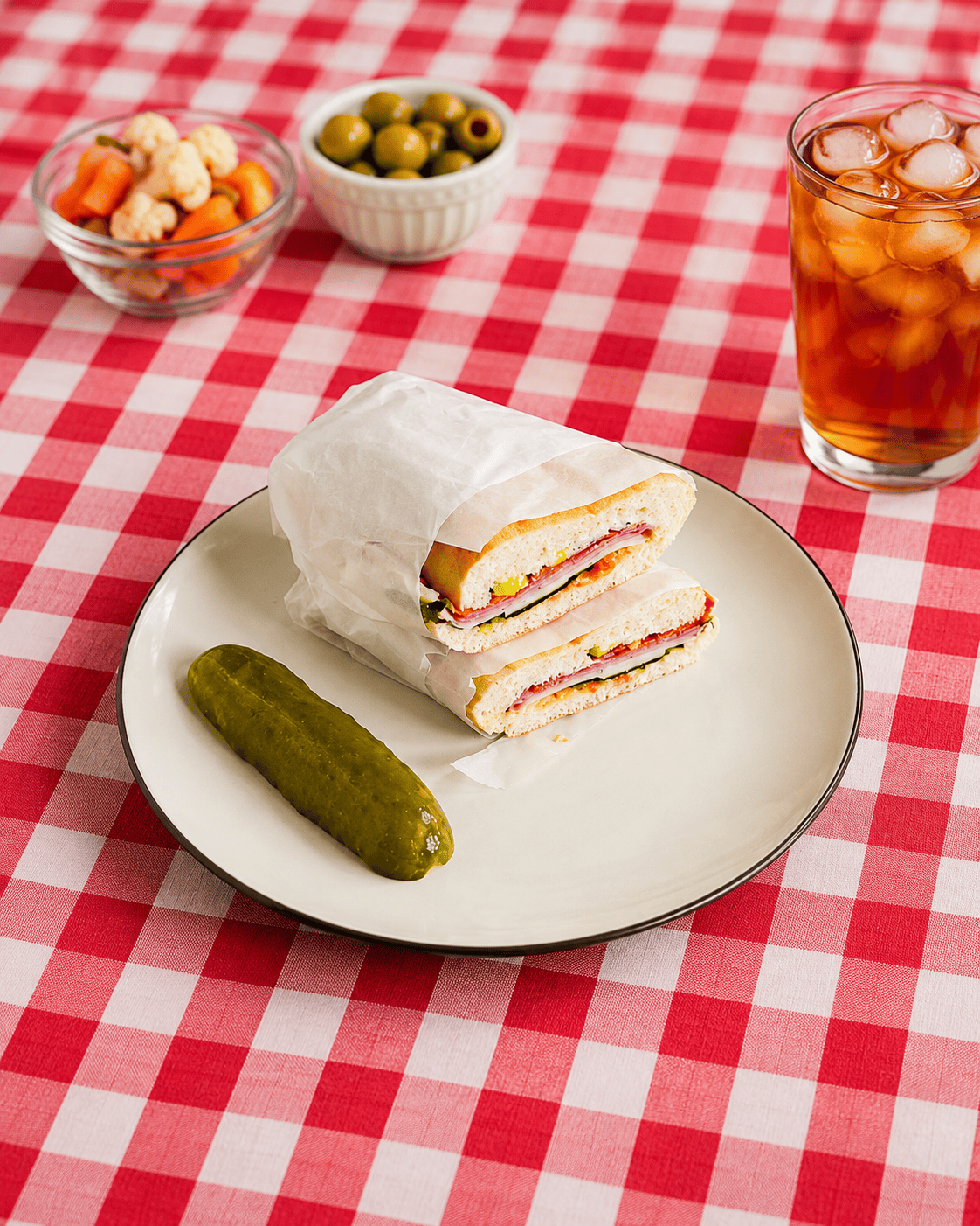 A plate with 2 sandwich halves stacked on top of each other and a pickle with a bowl of olives, a bowl of pickled veggies, and a glass of iced tea behind the plate.