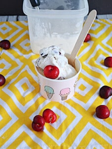 A cup of cherry vanilla ice cream with cherries all around and the container of ice cream in the background.