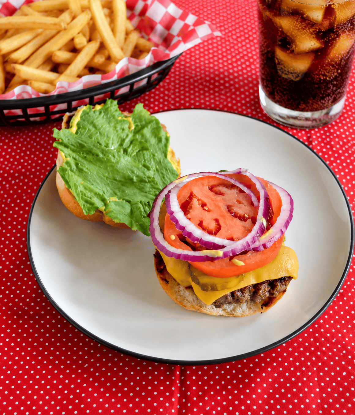 A plate with a smashburger on a bun with a basket of fries behind the plate and a glass of soda.