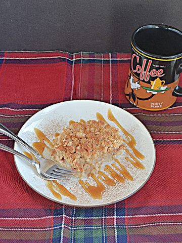A plate topped with a Pear Pie Bar drizzled with caramel sauce, two forks on the plate, and a coffee cup sitting behind it.