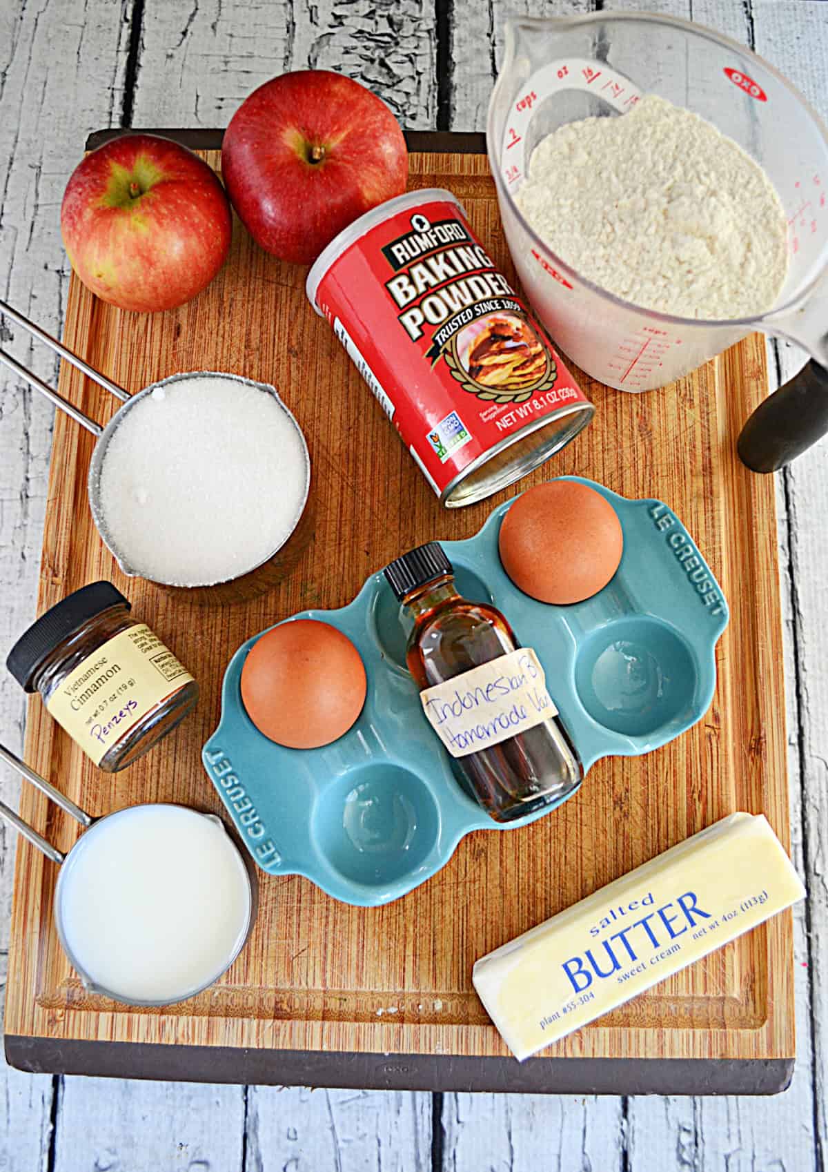 A cutting board with eggs, butter, a cup of sugar, a cup of flour, vanilla, 2 apples, and cinnamon on it.