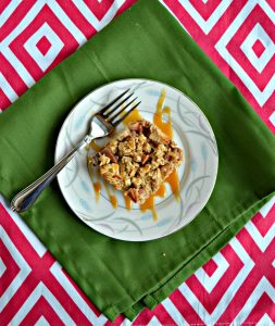 A white plate topped with an oatmeal square that is brown with apple pieces and has a caramel drizzle on top with a fork on the left side of the plate sitting on a green napkin on a red and white background.