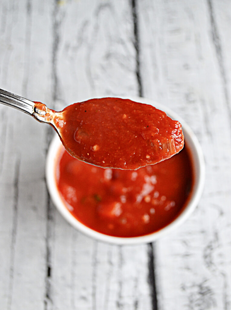 A spoon of tomato sauce with a bowl of the sauce behind it. 