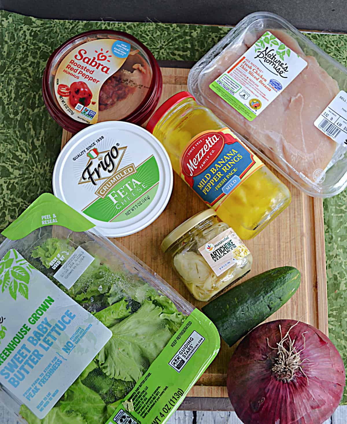 A cutting board with a bin of lettuce, feta cheese, a pack of chicken, a cucumber, a red onion, and a container of hummus on it.