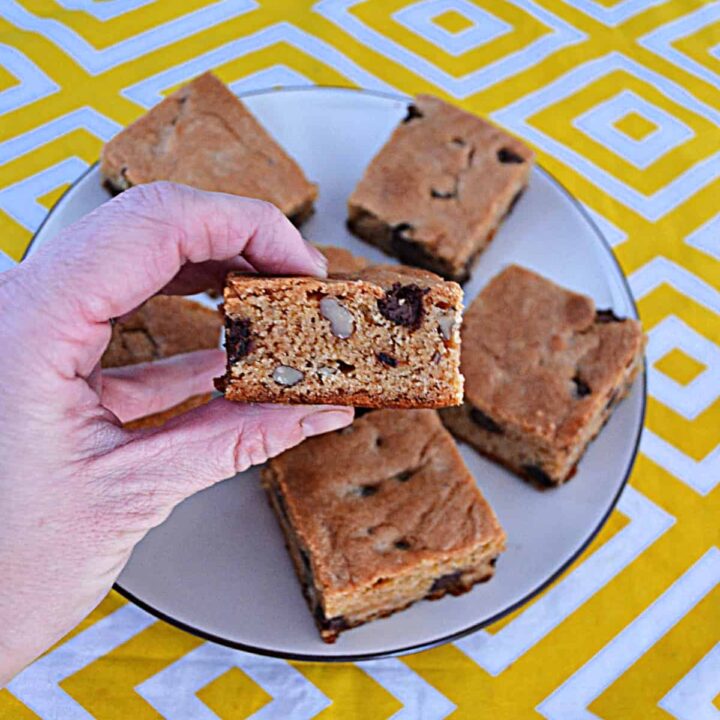 A hand holding a chocolate chip blondie with a plate of blondies behind it.