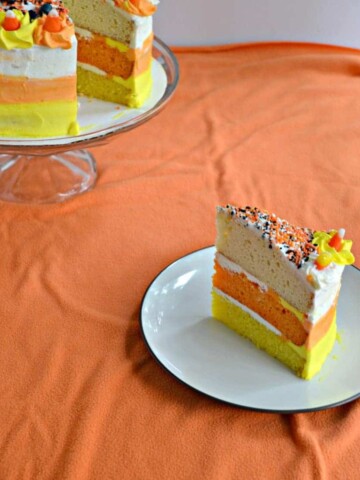 A slice of orange, yellow, and white candy corn cake with the whole cake behind it.