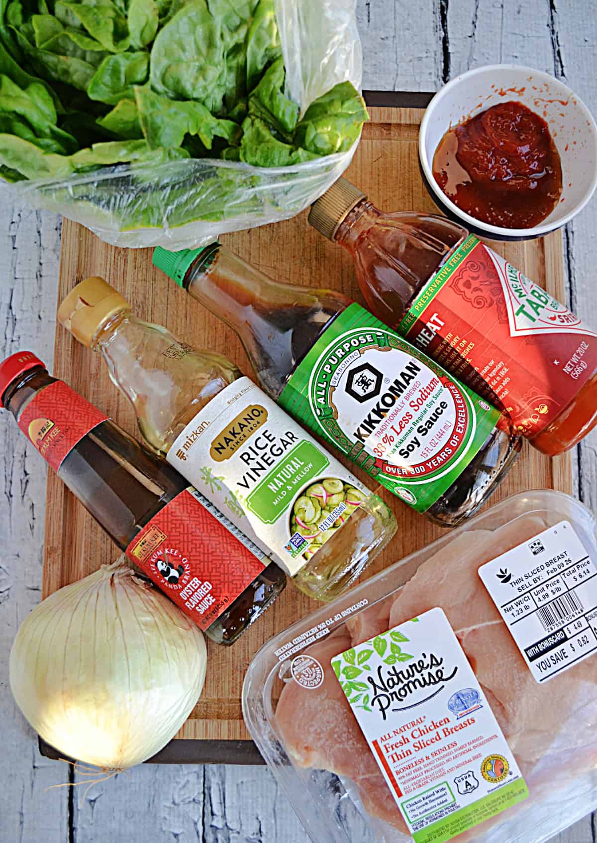 A cutting board with a head of lettuce, a bottle of soy sauce, rice vinegar, oyster sauce, an onion, a pack of chicken, and seasonings on it.