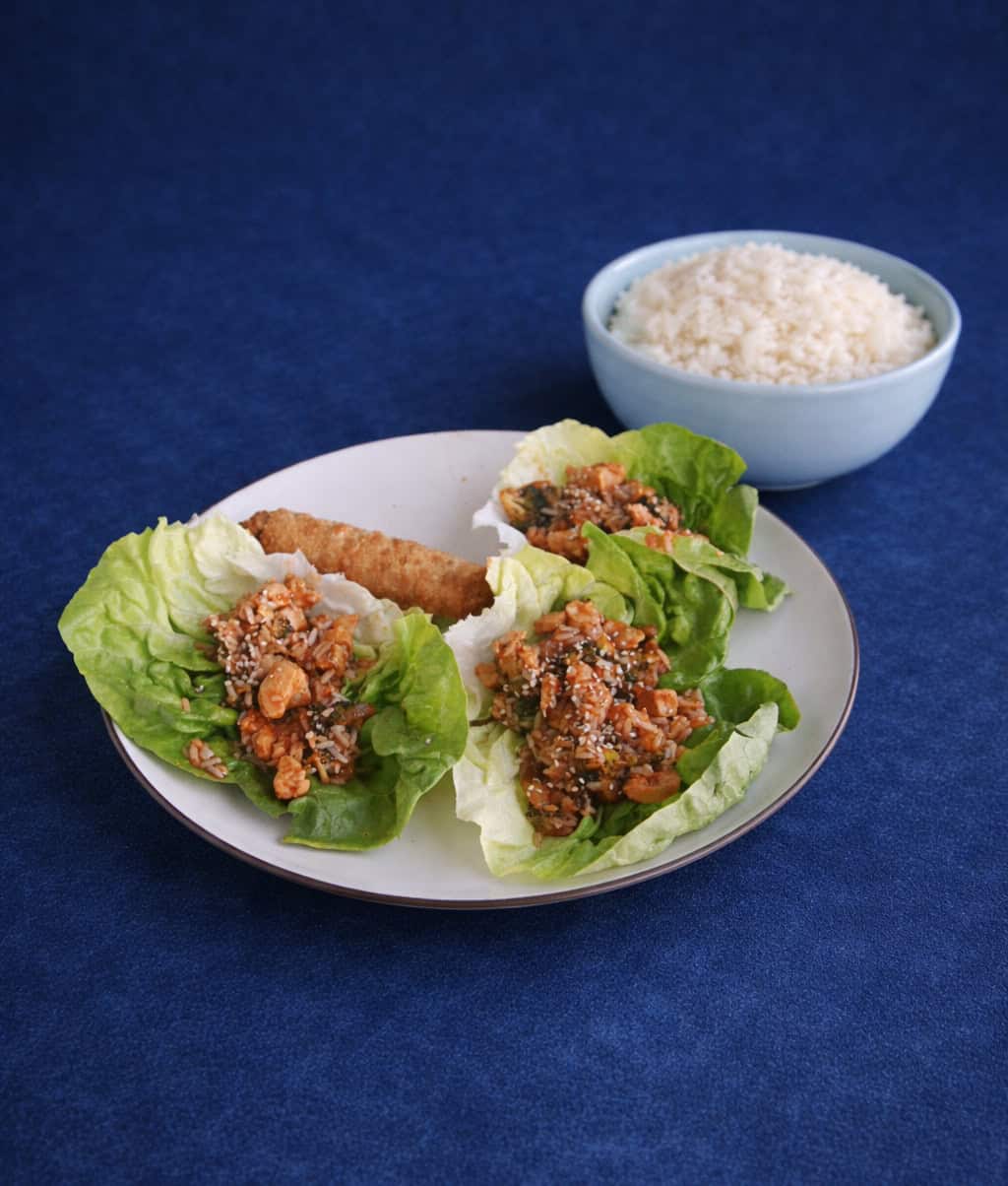 A plate with three lettuce wraps filled with chicken and an egg roll with a bowl of rice in the background.