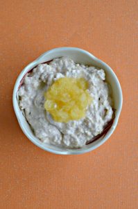 A bowl filled with cream cheese with a spoonful of rushed pineapple on top on an orange background.