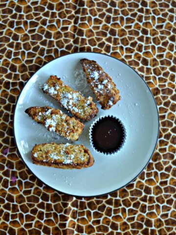 A plate with 4 French Toast sticks in a semi-circle sprinkled with powdered sugar with a cup of syrup on a cheetah background.