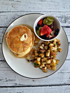 Close up of plate with a stack of pancakes, a pile of home fries, and a small bowl of fresh fruit.