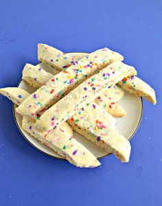 A blue background with a small plate on it topped with rainbow sprinkle biscotti