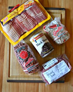 A cutting board with a package of bacon, 2 packages of dried beans, a can of black beans, and a package of ground beef.