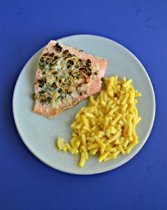 A piece of broiled salmon topped with herbs with a side of macaroni and cheese on a plate with a blue background.