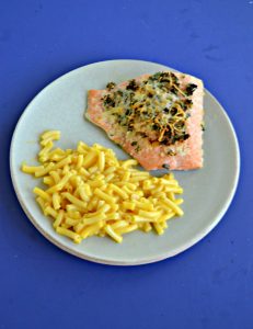 A plate with a large piece of salmon topped with herb and a side of macaroni and cheese on a blue background.