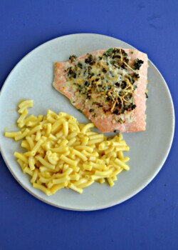 A plate with a large piece of salmon topped with herbs and a serving of macaroni and cheese with a blue background.
