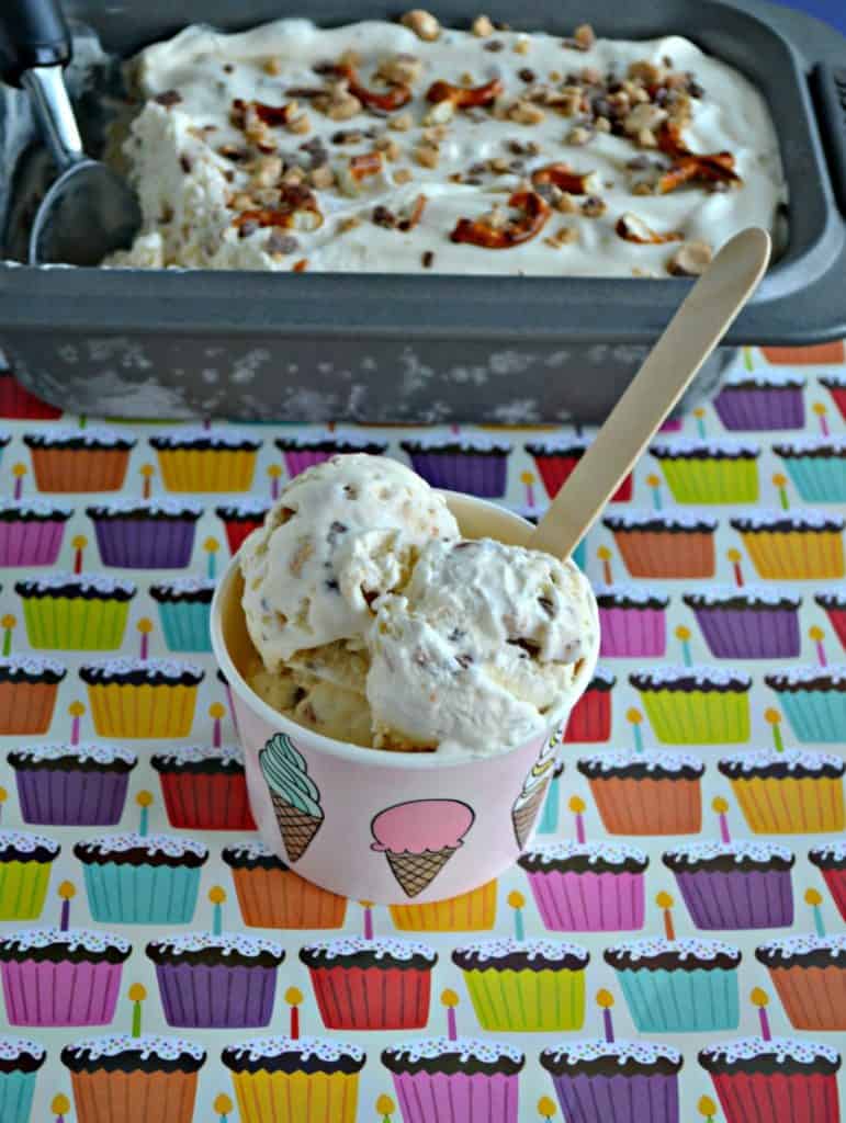 A cup of ice cream with 3 scoops and a wooden spoon with a loaf pan of ice cream behind it with an ice cream scoop in it.