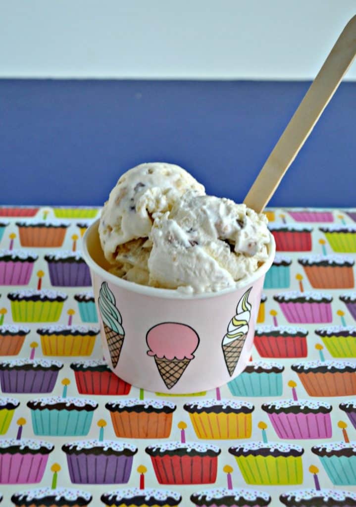 A cup of ice cream with 3 scoops and a wooden spoon sitting on a placemat with colorful cupcakes on it.