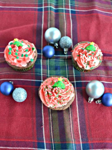 3 cupcakes with red frosting on a red plaid placemat with blue and silver ornaments