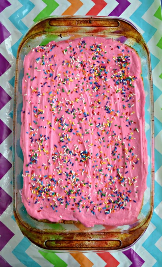 A pan of Funfetti Blondies topped with pink buttercream frosting and rainbow sprinkles on a rainbow background.