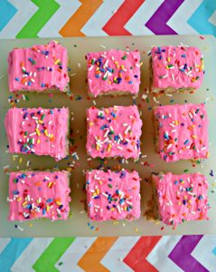 Three rows of blondies with three in each row. Each topped with bright pink frosting and rainbow sprinkles on a white cutting board on a rainbow tablecloth.