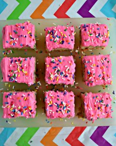 Three rows of blondies with three in each row. Each topped with bright pink frosting and rainbow sprinkles on a white cutting board on a rainbow tablecloth