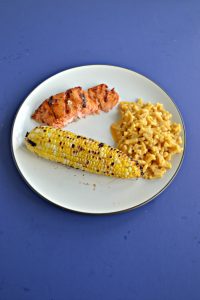 A white plate with a piece of grilled corn, a spoonful of macaroni and cheese, and a grilled salmon filet on a blue background.