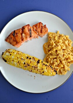 A white plate with an ear of grilled corn, a spoonful of macaroni and cheese, and a grilled salmon filet.