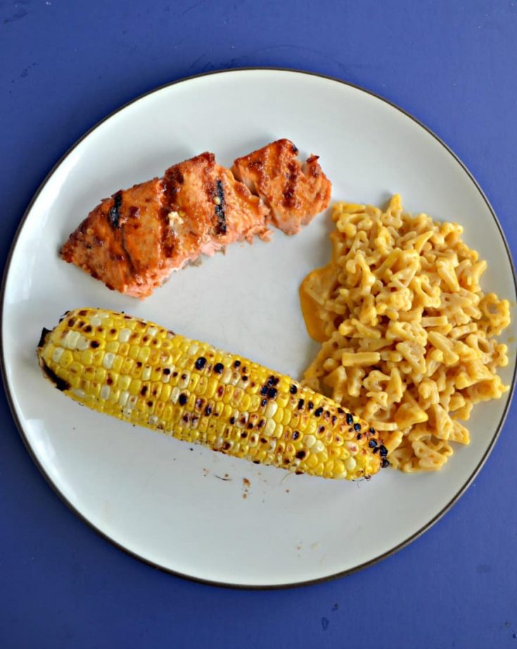 A white plate with an ear of grilled corn, a spoonful of macaroni and cheese, and a grilled salmon filet.