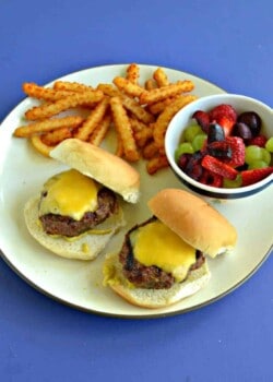 A plate with two cheeseburger sliders, a handful of fries, and a bowl of mixed fruit and berries on a blue background.