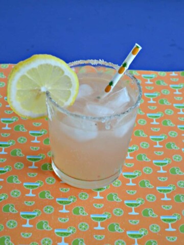 Short glass with a margarita on the rocks with a lemon slice on an orange and green margarita backdrop.