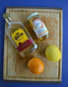 A bottle of tequila, a jar of honey, an orange, and a lemon on a wooden cutting board.