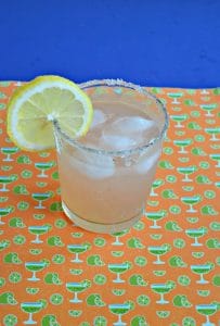 Glass of margarita on the rocks with a slice of lemon on an orange and green backdrop.