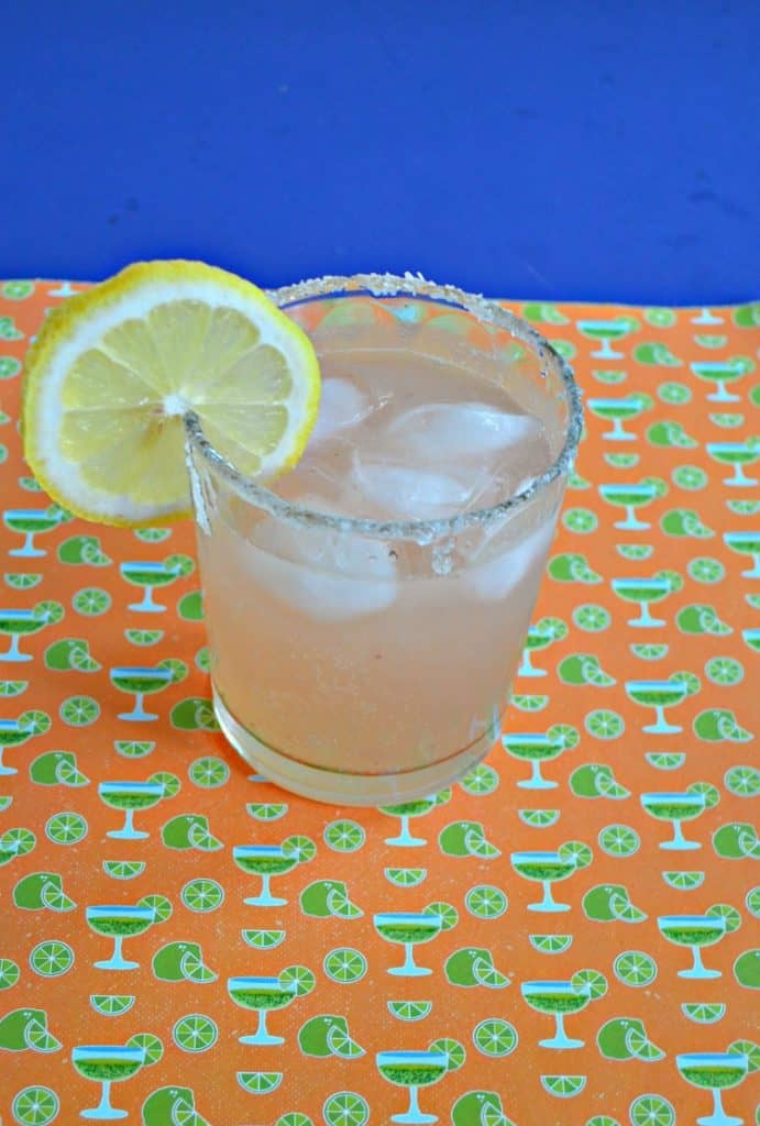 Glass of margarita on the rocks with a slice of lemon on an orange and green backdrop.