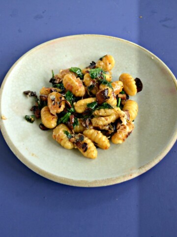 A plate mounded with browned gnocchi, wilted spinach, and mushrooms on a blue background.