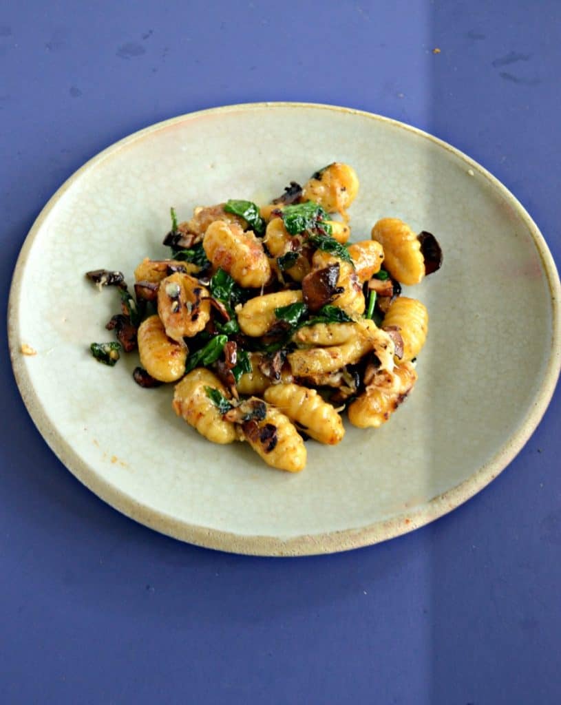 A plate mounded with browned gnocchi, wilted spinach, and mushrooms on a blue background.
