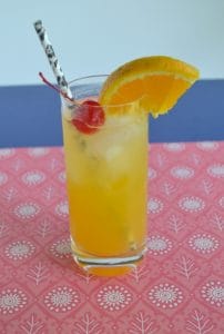 Black Eyed Susan: Tall glass with orange drink in it topped off with a cherry, orange slice, and black straw, sitting on a red placemat with a blue background.