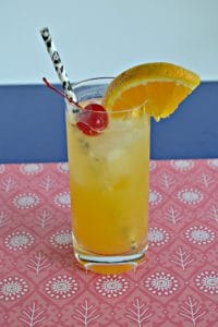 Tall glass with orange drink in it topped off with a cherry, orange slice, and black straw, sitting on a red placemat with a blue background.