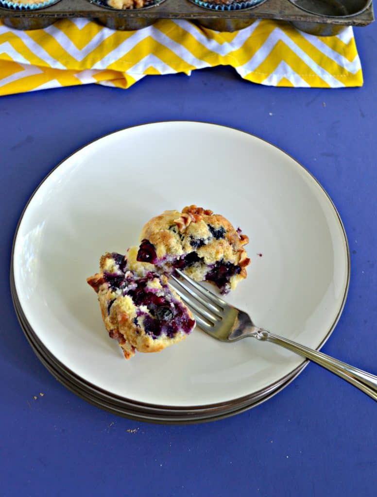 A blueberry muffin on a plate that is split open with a fork sitting in between the pieces and a yellow napkin in the background.