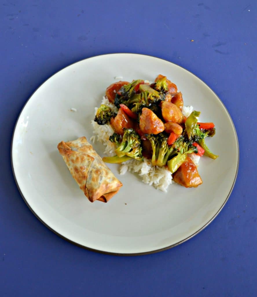 A plate with an egg roll on one side and a pile of rice topped with chicken, broccoli, and red peppers on the other side on a blue background.