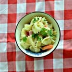 A bowl of bow tie pasta with fresh herbs, deli meats, and carrots on a red and white checked background