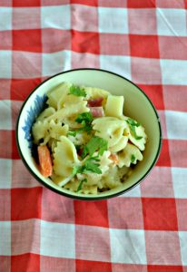 Close up view of a bowl of bow tie pasta with fresh herbs, meats, and veggies on a red and white checked background.