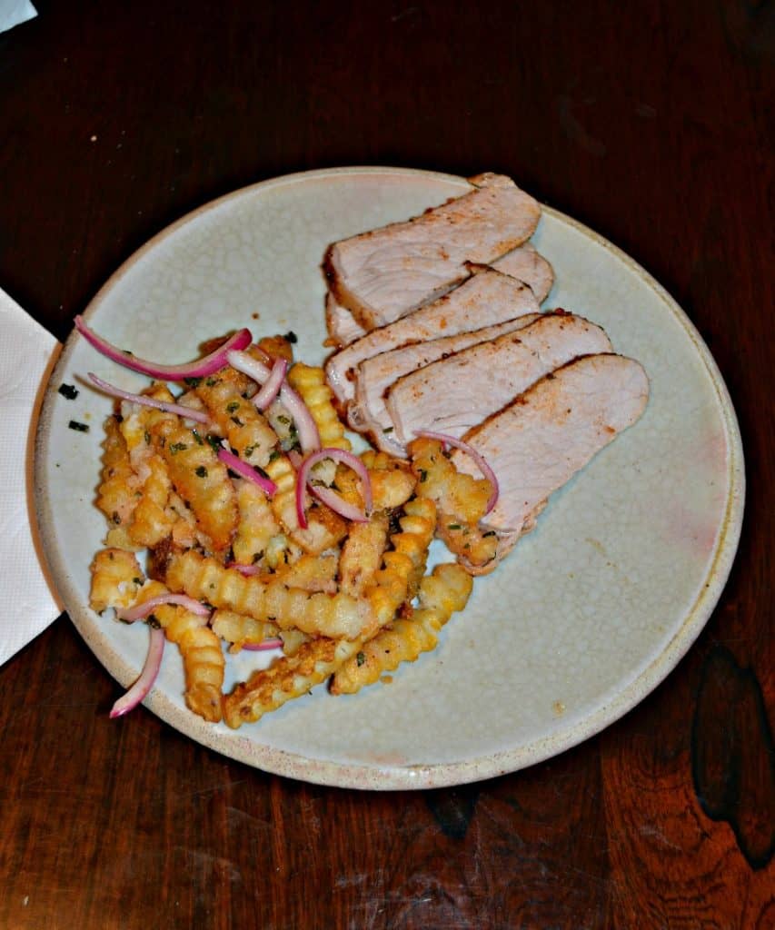 A plate with 5 slices of pork loin and a pile of crinkle cut fries with red onions and chives.