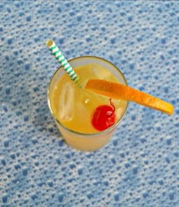 Top view of cocktail: Orange liquid with blue and white stripe straw sticking up, a bright red cherry, and an orange slice with a blue bubble background.