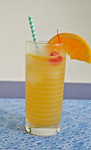 Close up view of orange cocktail with blue and white striped straw, orange slice, bright red cherry, on a blue bubble placemat.
