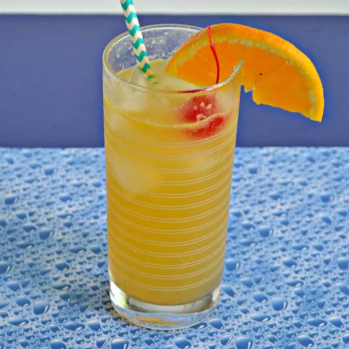Side view of cocktail: Orange liquid with blue and white stripe straw sticking up, a bright red cherry, and an orange slice with a blue bubble background.