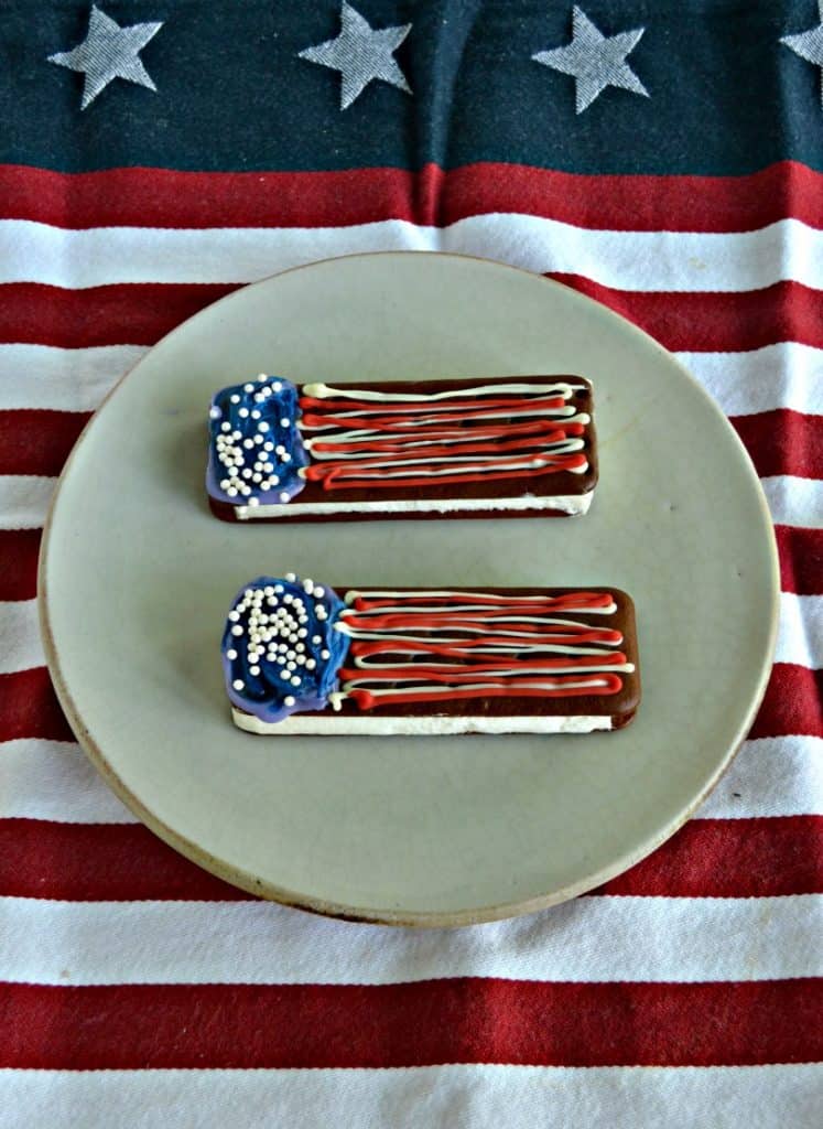 Two ice cream sandwiches decorated with blue chocolate on the left with edible pearls and red and white horizontal squiggles on nthe right to resemble a flag. Sitting on a red and white tablecloth.