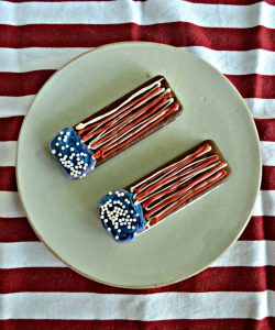 Two ice cream sandwiches sitting tilted on a plate decorated with blue chocolate on the left with edible pearls and red and white horizontal squiggles on nthe right to resemble a flag. Sitting on a red and white tablecloth.