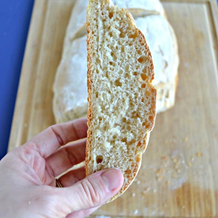 A close up look of a slice of sourdough bread being held up in front of a sourdough bread loaf.